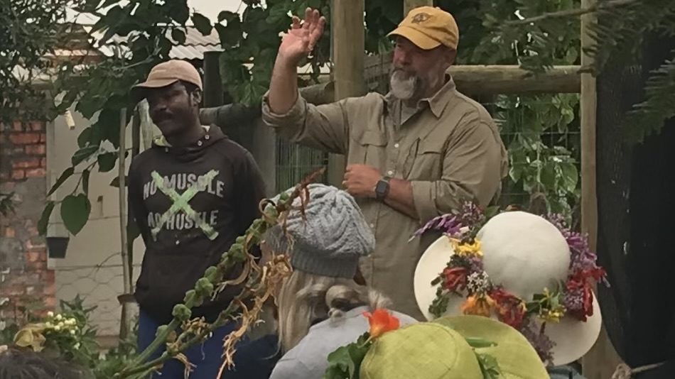 Two men in caps speak to a group of people, seen from behind, in a garden with a trellis and green foliage